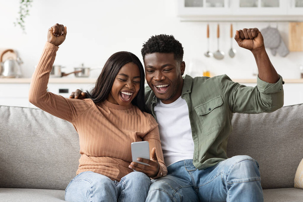 Happy black lovers using smartphone together and gesturing, home interior - Zdjęcie, obraz