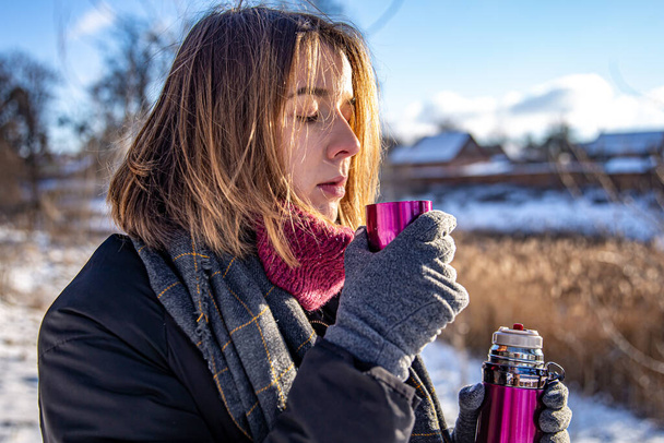 Μια νεαρή γυναίκα απολαμβάνει ένα ζεστό ποτό από ένα θερμός σε μια βόλτα το χειμώνα. - Φωτογραφία, εικόνα