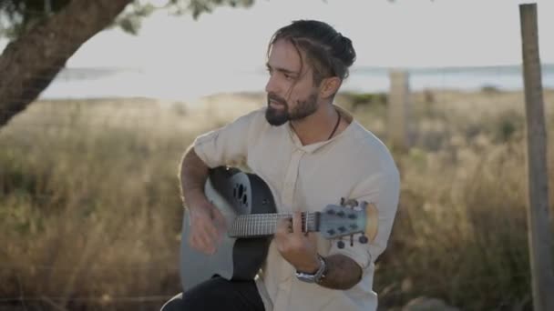 Knappe man gitarist op klassieke akoestische gitaar en zanglied kijkend naar cameralens op het platteland van Spanje bij zonsondergang  - Video