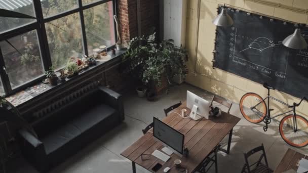 Wysoki kąt widzenia nowoczesnej przestrzeni współpracowników ze stołami, komputerami, sofą, zielonymi roślinami, tablicą i rowerem na ścianie w słoneczny letni dzień - Materiał filmowy, wideo