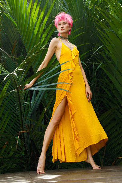 Модница с розовыми умирающими волосами надевает желтое платье и выглядит сильной над зелеными листьями. Азиатская тонкая женщина элегантно позиционирует себя в качестве модной съемки в концептуальной стране летней тропической зоны - Фото, изображение