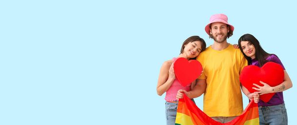 Hombre y dos hermosas mujeres con bandera de arco iris y corazones rojos sobre fondo claro con espacio para el texto. Concepto de poliamory y LGBT - Foto, imagen