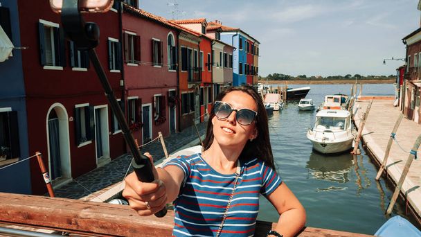 メディアに影響を与えるイタリアのブラーノ。ヨーロッパヴェネツィア市内のペイントハウス。ヴェネツィア島のカラーストリートでスマートフォンで旅行写真家ブロガーの女の子。自由、夏の旅行、職業概念 - 写真・画像