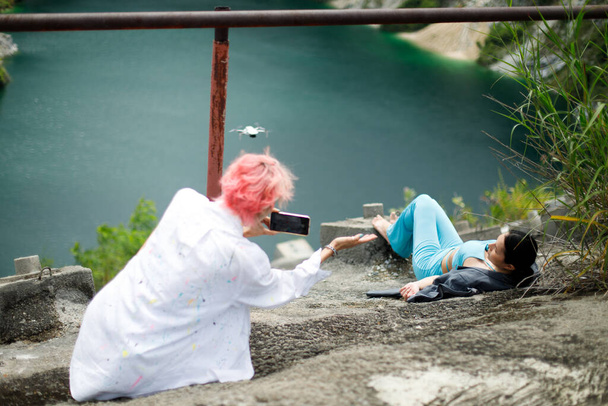 Asyalı Kadın Fotoğrafçı, cep telefonu kamerasını, yalancı kadını uçurumdan aşağı çekmek için tut. Moda mankeni dağ gölü manzarasında oturuyor. Sosyal medya için fotoğraf çekimi var ama tehlikeli bir yerde. - Fotoğraf, Görsel