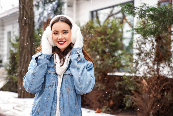 Beautiful young woman wearing mittens and earmuffs on snowy day - Photo, Image