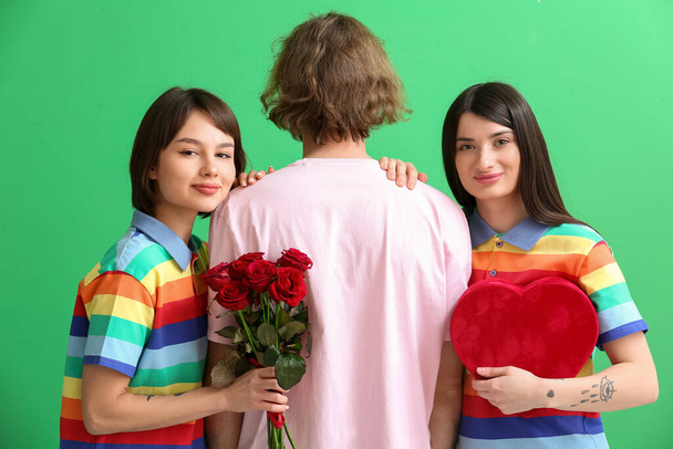 Mężczyzna i dwie piękne kobiety w tęczowe ubrania, z prezentem i kwiatami na tle koloru. Koncepcja poliamii i LGBT - Zdjęcie, obraz
