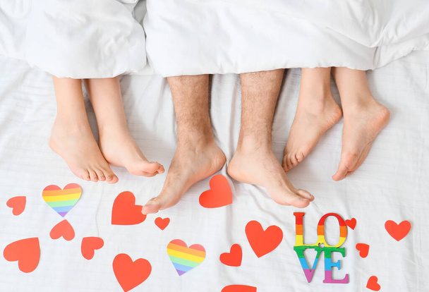 Piernas de hombre joven y dos mujeres tumbadas en la cama cubiertas de corazones de papel, vista superior. Concepto de poliamory y LGBT - Foto, Imagen