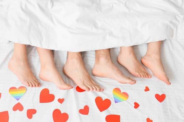 Piernas de hombre joven y dos mujeres tumbadas en la cama cubiertas de corazones de papel, vista superior. Concepto de poliamory y LGBT - Foto, imagen