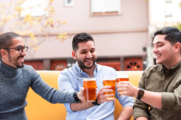Grupa latynoskich przyjaciół, szczęśliwa, wznosząca toast i uśmiechnięta, pijąca piwo rzemieślnicze, siedząca na żółtej kanapie, przed barem. koncepcja stylu życia. - Zdjęcie, obraz