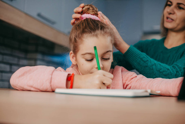 Caucasian girl writing in her notebook while her mother makes her look good. Mother helping daughter homework, great design for any purposes. Children education - Фото, изображение