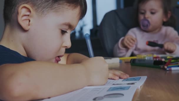 宿題をしている間にコピー本でノートを作る彼の妹とテーブルに座っている男の子とのビデオ。子供教育。距離教育. - 映像、動画