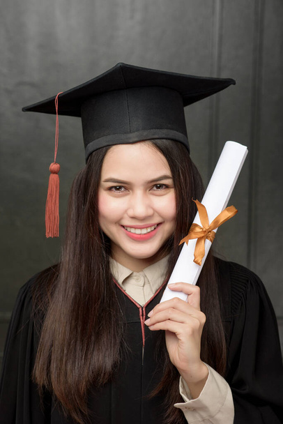 Portrait of young woman in graduation gown smiling and cheering on black background  - Foto, Imagem
