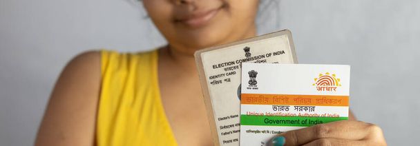 Birbhum, Bengala Occidental / India - 21 de diciembre de 2021: Enfoque selectivo en Aadhaar y tarjeta de votante de la India sostenida en la mano por una joven india con rostro sonriente - Foto, imagen
