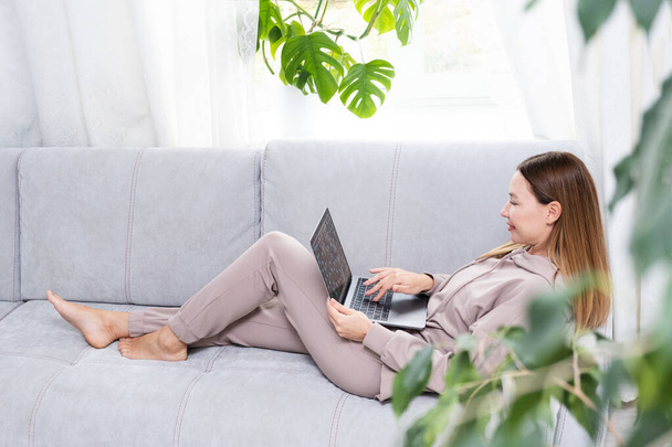 Mujer joven o de mediana edad sentada con portátil en sofá gris en la oficina en casa con planta de monstera. Concepto de lugar de trabajo remoto y trabajo en el hogar. - Foto, Imagen