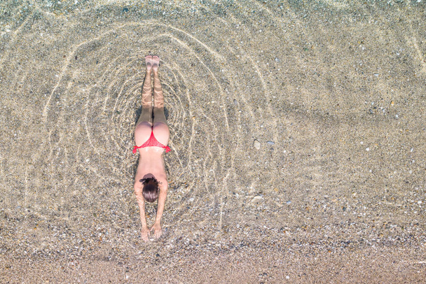 Top, aerial view. Young beautiful woman in a red bikini panties lying and sunbathe in sea water on the sand beach. Drone, copter photo. Summer vacation. View from above.  - Valokuva, kuva