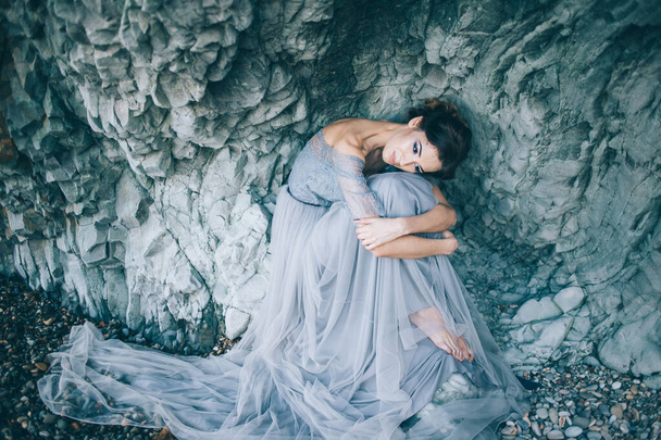 Beautiful girl bride in a long blue dress sitting near the cliff bowed his head, hugging his knees - Foto, Imagen