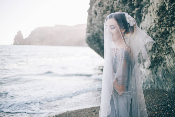 Beautiful brunette girl bride in a grey gown of lace and tulle, covered her face with a veil, hand on breast, profile, horizontal, sea, waves, summer - Φωτογραφία, εικόνα