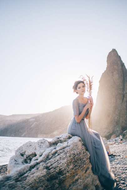 Beautiful brunette woman in long dress sitting white stone by the sea at sunset and looking at the camera. Beauty, wedding, fine art concept - Φωτογραφία, εικόνα