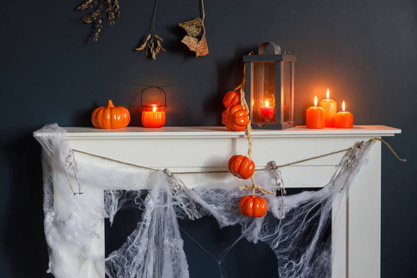 Halloween home decoration. Plastic toy skeletons in a wooden box on the fireplace against a dark blue wall. A garland of skeletons. Cobweb on the dresser. Orange candles and lantern. - 写真・画像