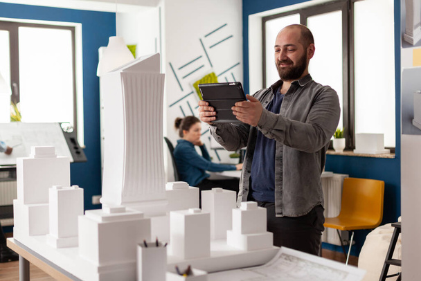 Bearded professional architect looking at tablet in front of white foam building model - Photo, image