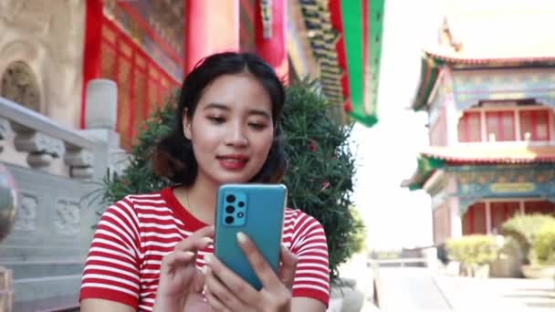 Portrait asian woman wearing a red shirt visits a temple on Chinese New Year, sits in a chair, plays with her smartphone and looks at the dial with a cheerful, amused face. - Footage, Video