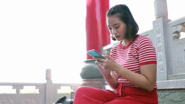 Young asian woman in bright red clothes sits on a smartphone before looking at the camera and smiling, good-natured. - Footage, Video