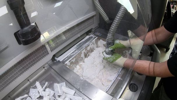 Человек очищает детали, напечатанные на промышленном 3D принтере из белого пластикового порошка - Фото, изображение