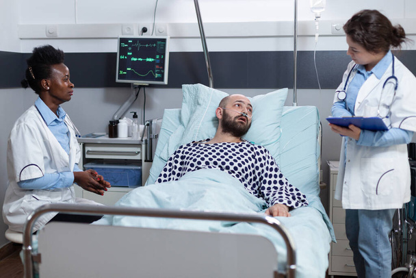 Γιατρός κρατώντας πρόχειρο με κλινικά δεδομένα συμβουλεύεται την ανάκτηση του ασθενούς, ενώ οι μόνιμοι κάτοικοι παρακολουθούν ζωτικά όργανα - Φωτογραφία, εικόνα