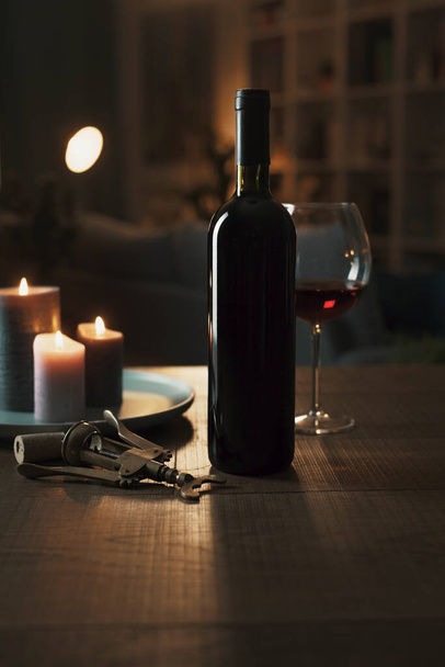 Red wine tasting at home: wine bottle, wineglass, corkscrew and candles on a table in the living room at night - Foto, Imagen