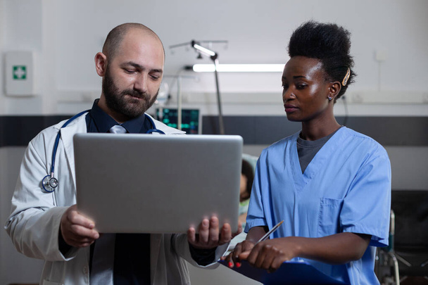Arts holding laptop presenteert patiënt medische geschiedenis aan verpleegkundige holding klembord met laboratoriumresultaten - Foto, afbeelding