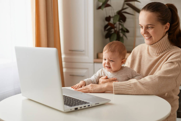 Indoor shot of smiling happy woman with ponytail hairstyle wearing casual style beige sweater sitting at table in kitchen with her toddler baby and looking at laptop display with positive expression. - Foto, Imagen