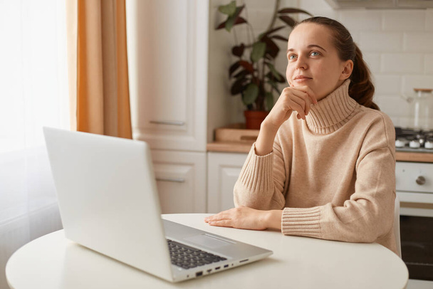 Portrait of pensive woman with ponytail hairstyle wearing casual style beige sweater sitting at table in kitchen and working on computer, dreaming about rest, looking away and holding chin. - Foto, afbeelding