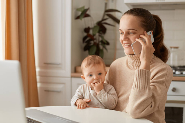 Indoor shot of happy smiling woman wearing beige casual style sweater with ponytail hairstyle sitting at table with her toddler daughter and talking phone with optimistic facial expression. - Фото, зображення