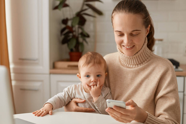 Portrait of woman wearing beige casual style sweater with ponytail hairstyle sitting at table with her infant daughter and using smart phone with positive facial expression. - Valokuva, kuva