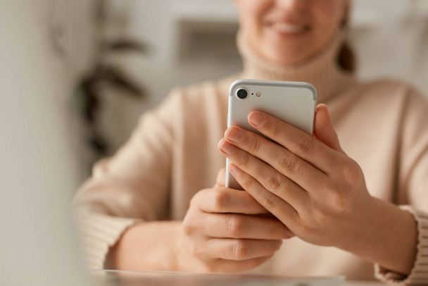 Closeup portrait of woman wearing beige casual style sweater using cell phone, watching videos in social networks or typing e-mail, expressing positive emotions, smiling. - 写真・画像