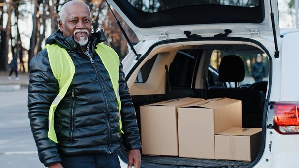 制服を着た高齢アフリカ系アメリカ人男性の宅配業者は、労働者が積極的に仕事をして配達を取る特別な服で車の便利屋ローダーから段ボール箱をアンロードする屋外で成熟した商品を取ります - 写真・画像
