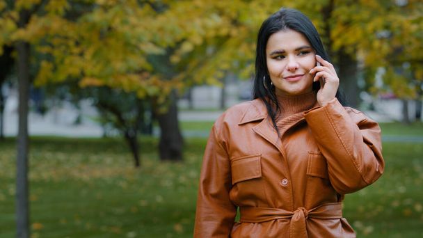 Young smiling woman outdoors in autumn park talking on mobile phone hispanic girl enjoys pleasant conversation communicates by telephone female customer makes order in online store using smartphone - Zdjęcie, obraz