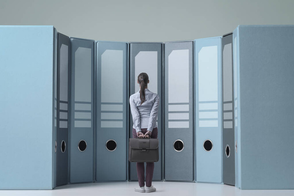 Confused businesswoman standing in front of huge ring binders in the archive, data management and storage concept - 写真・画像