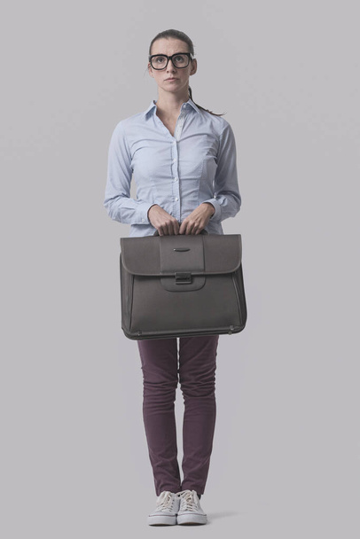 Young shy businesswoman standing and holding her briefcase, isolated on gray background - Valokuva, kuva