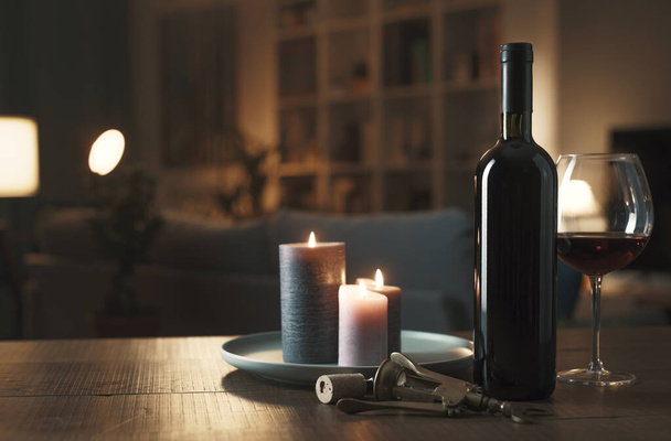 Excellent red wine tasting: bottle, wineglass, lit candles and corkscrew on a table in the living room - Фото, зображення