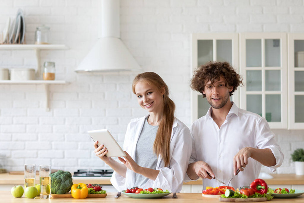 Portrait of a cheerful young couple cooking salad together according to a recipe on a tablet computer, looking at camera. Healthy eating. - Photo, image