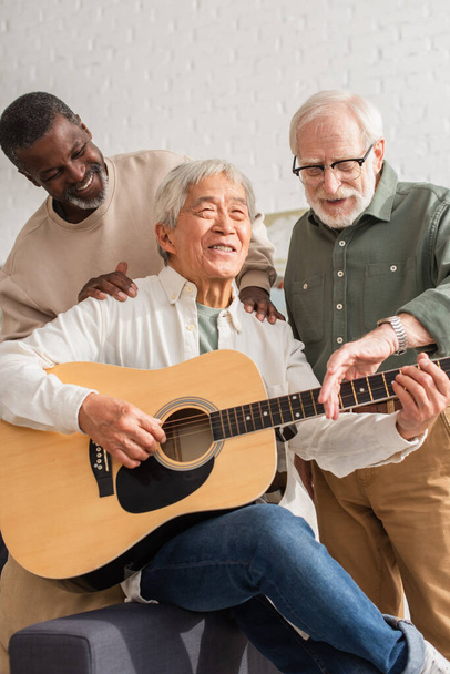 feliz sénior multiétnico hombres de pie cerca asiático amigo jugando acústica guitarra en casa  - Foto, Imagen