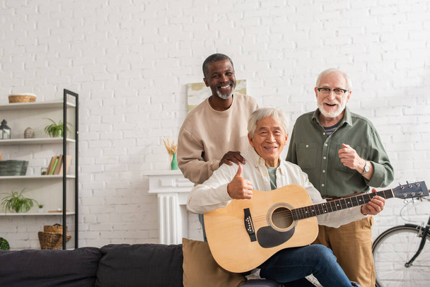Smiling asian man holding acoustic guitar and showing like near interracial friends at home  - Foto, afbeelding