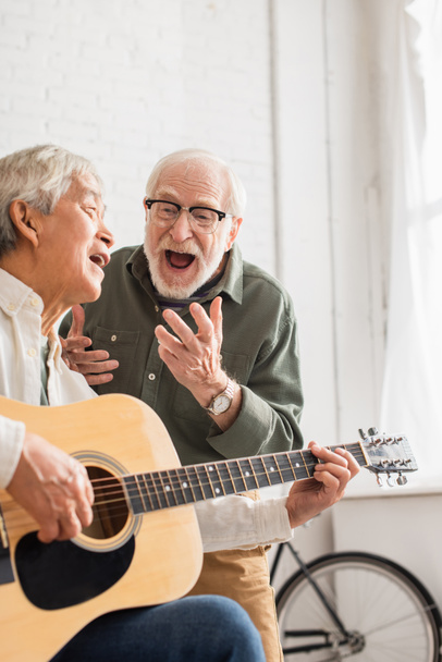 Podekscytowany starszy mężczyzna śpiewa w pobliżu azjatyckiego przyjaciela z gitarą akustyczną w domu  - Zdjęcie, obraz