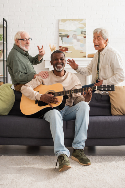 Interracial senior men singing near african american friend playing acoustic guitar at home  - Foto, afbeelding