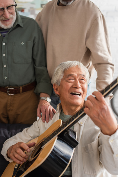 Yaşlı Asyalı adam evdeki ırklar arası arkadaşlarının yanında akustik gitar çalıyor.  - Fotoğraf, Görsel
