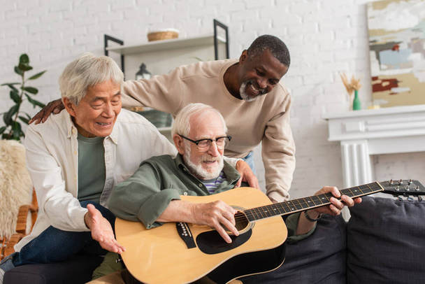 Positivos pensionistas interracial mirando a un amigo tocando la guitarra acústica en casa  - Foto, imagen