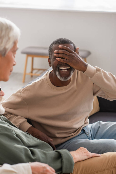 Cheerful african american man covering face near senior friends on couch  - Photo, Image