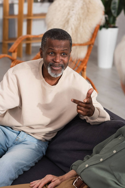 Smiling african american man talking to blurred elderly friends on couch  - Photo, image