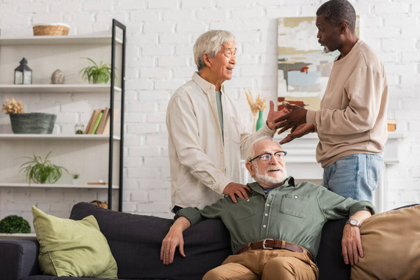 Multiethnic men talking near senior friend on couch at home  - Foto, Imagem